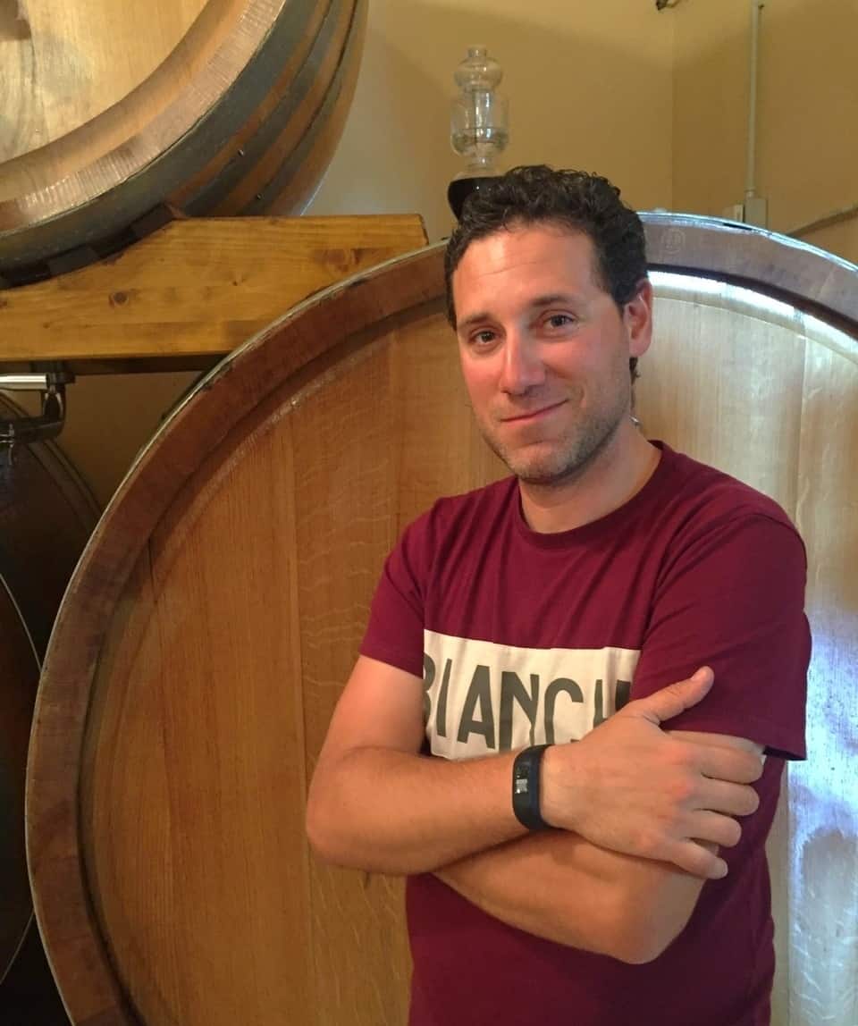 A man in a maroon t-shirt stands with arms crossed in front of large wooden barrels, perhaps pausing during one of his cycling tours or bike tours in Italy.