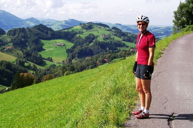 A cyclist wearing a helmet and red jersey stands on a roadside with rolling green hills and scattered houses in the background, capturing the scenic charm of bike tours in Italy.
