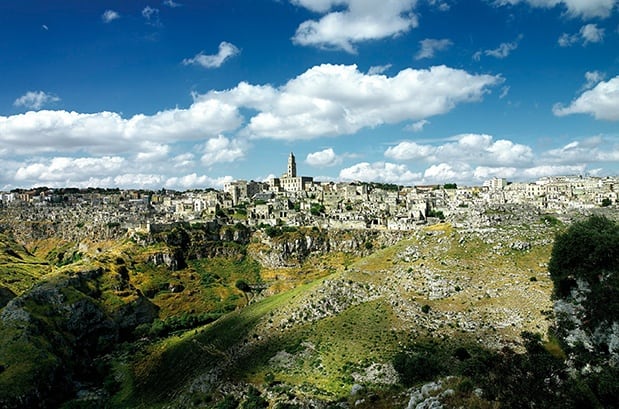 A panoramic view of an ancient stone city on a hilltop with rugged terrain and a deep ravine in the foreground under a partly cloudy sky, perfect for unforgettable bike tours in Italy.