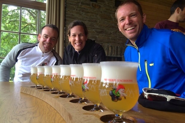 Three people sit at a wooden table smiling behind a row of filled beer glasses—celebrating the end of one of their international bike tours—with a window and stone wall in the background.
