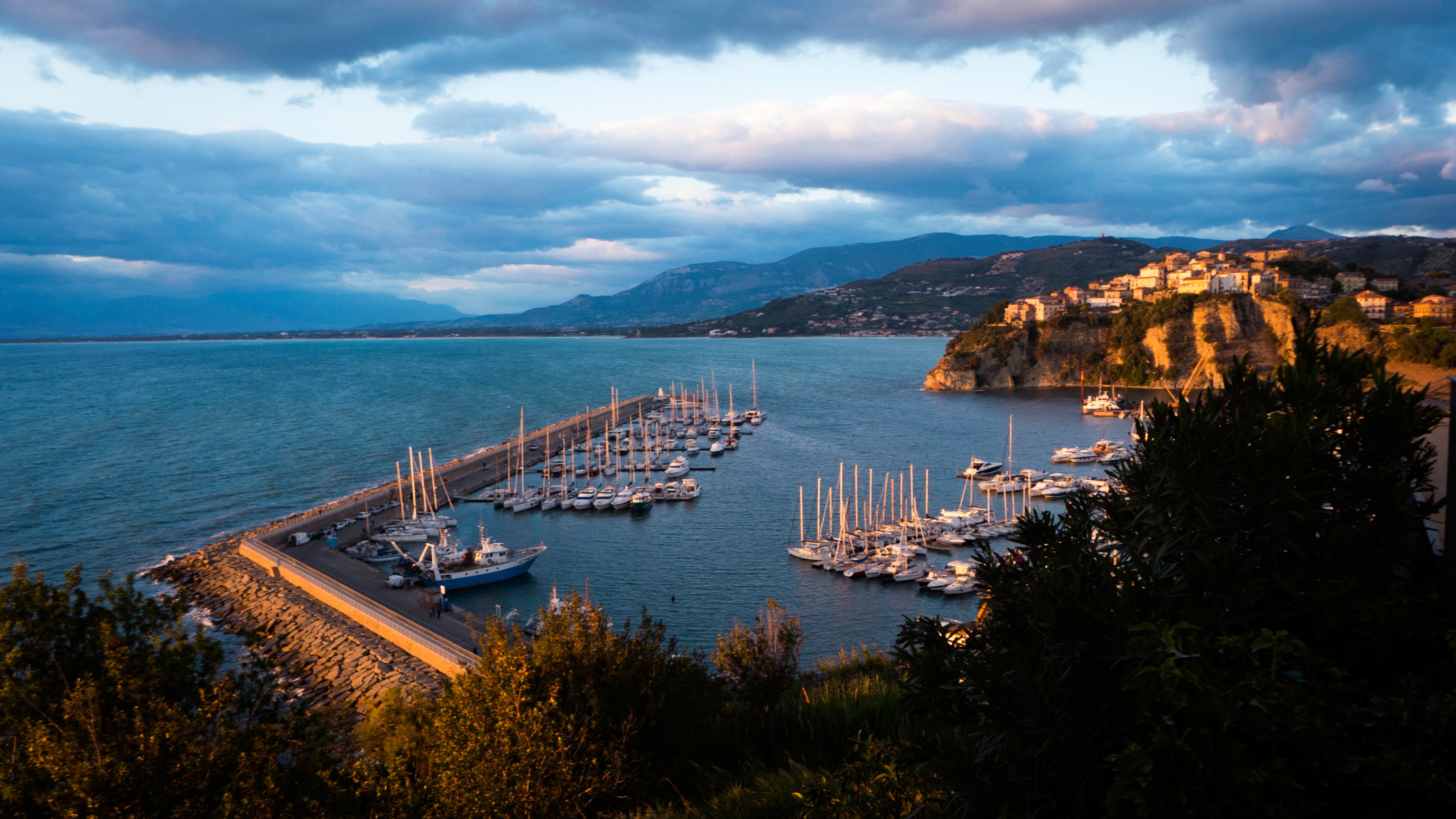 A marina with numerous sailboats docked, bordered by a breakwater, with a coastal town on a hill and mountains in the background under a cloudy sky—a perfect stop for scenic bike tours along the Italian coast.