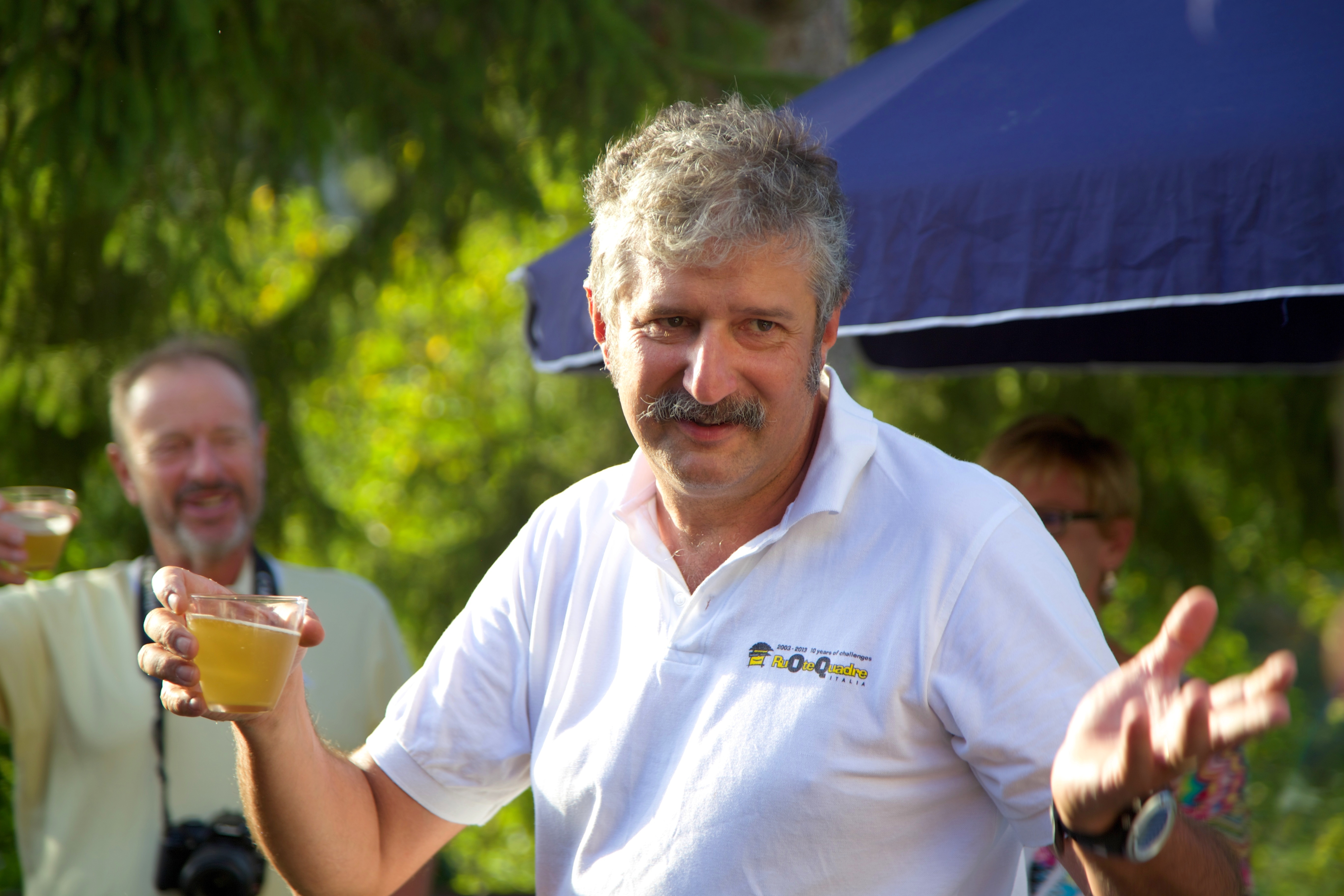 A man with a mustache holds a drink and gestures with his other hand outdoors, enjoying a break during one of the scenic cycling tours. Other people stand in the background, soaking in the atmosphere often found on bike tours in Italy.