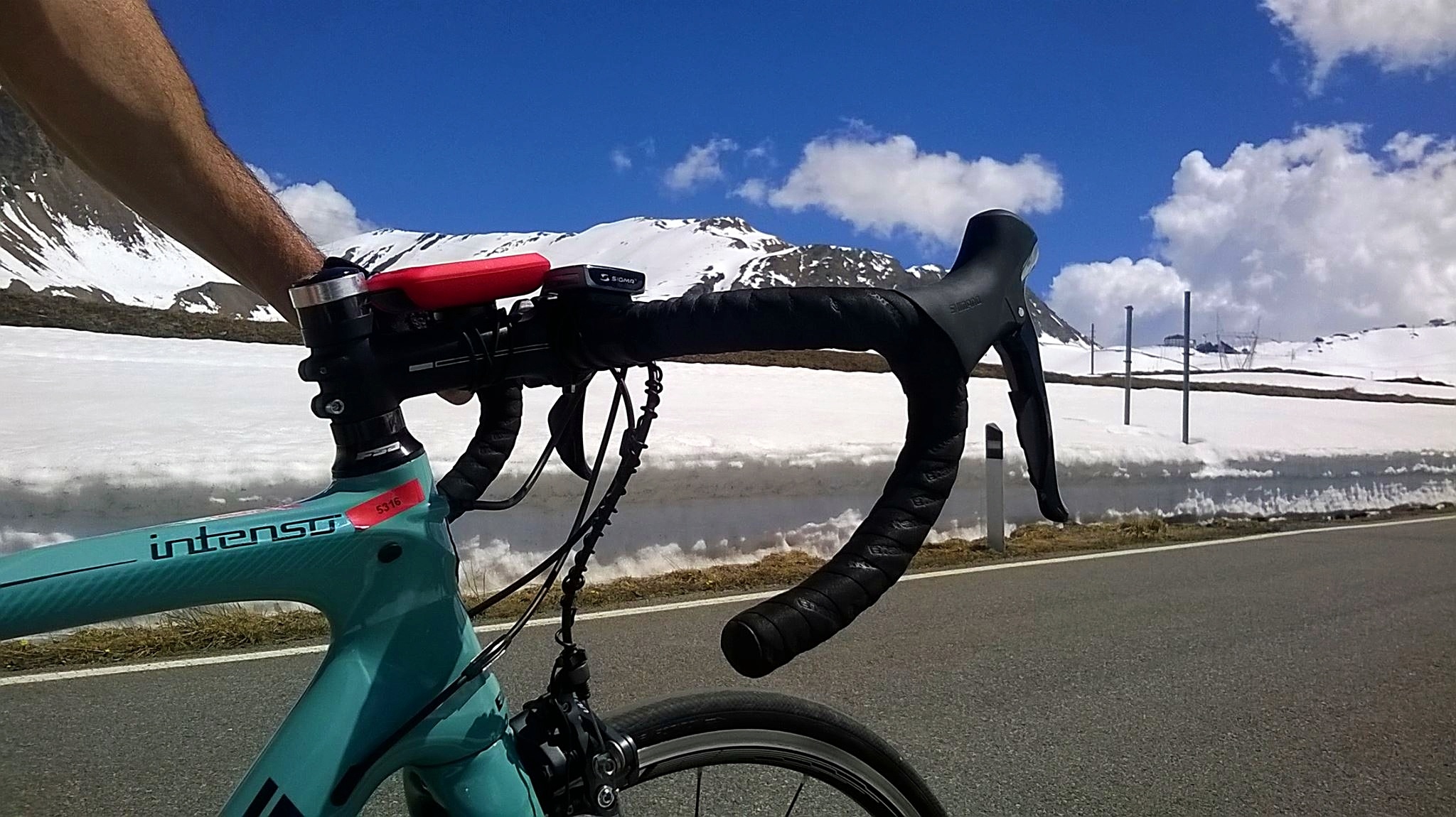 A person rides a teal road bike on a paved road beside snow-covered mountains under a blue sky with clouds, evoking the adventure and beauty found on cycling tours around the world.
