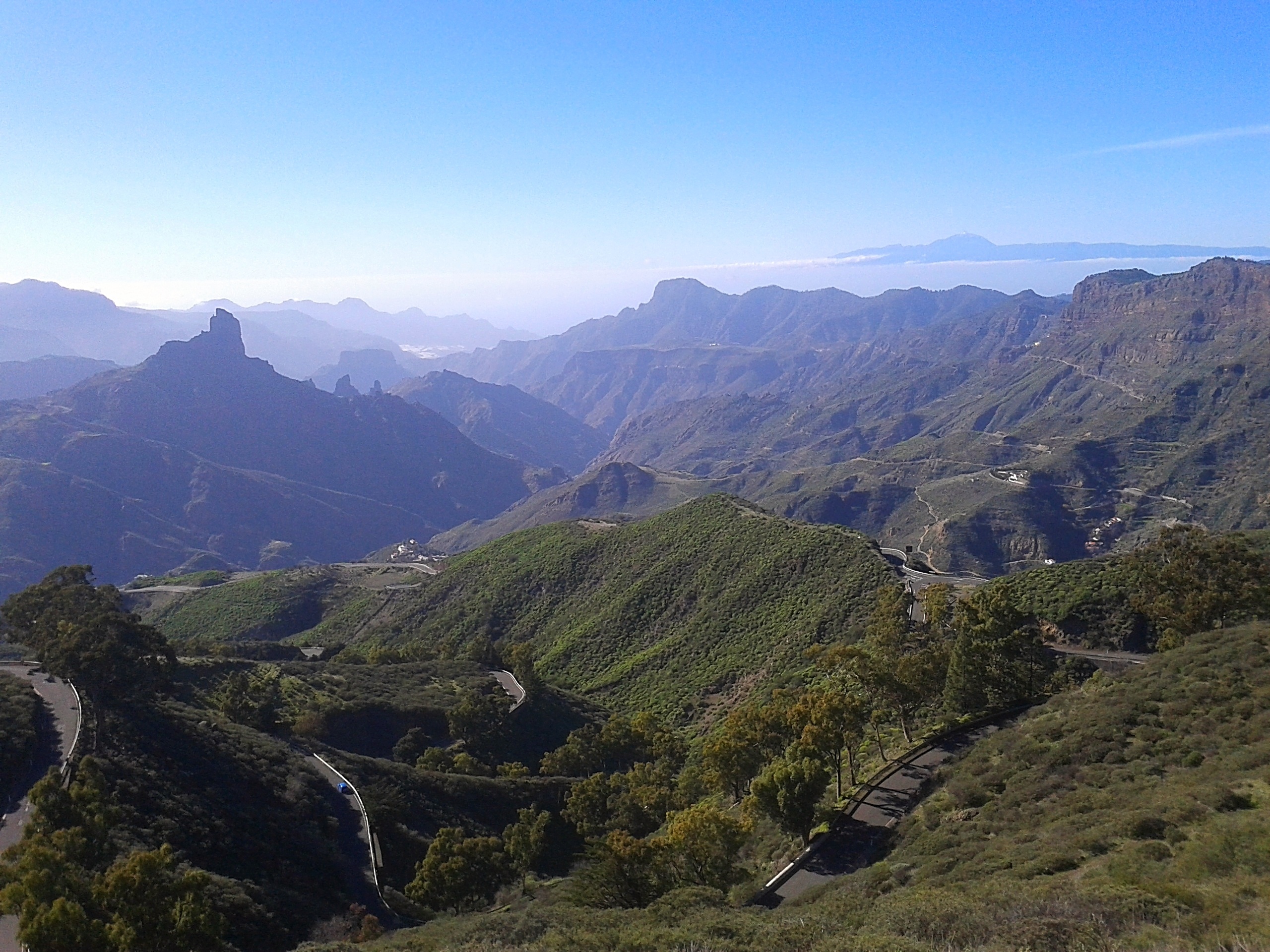 A winding road cuts through green hills and rugged mountains under a clear blue sky, offering breathtaking views perfect for scenic bike tours in Italy, with distant peaks visible on the horizon.