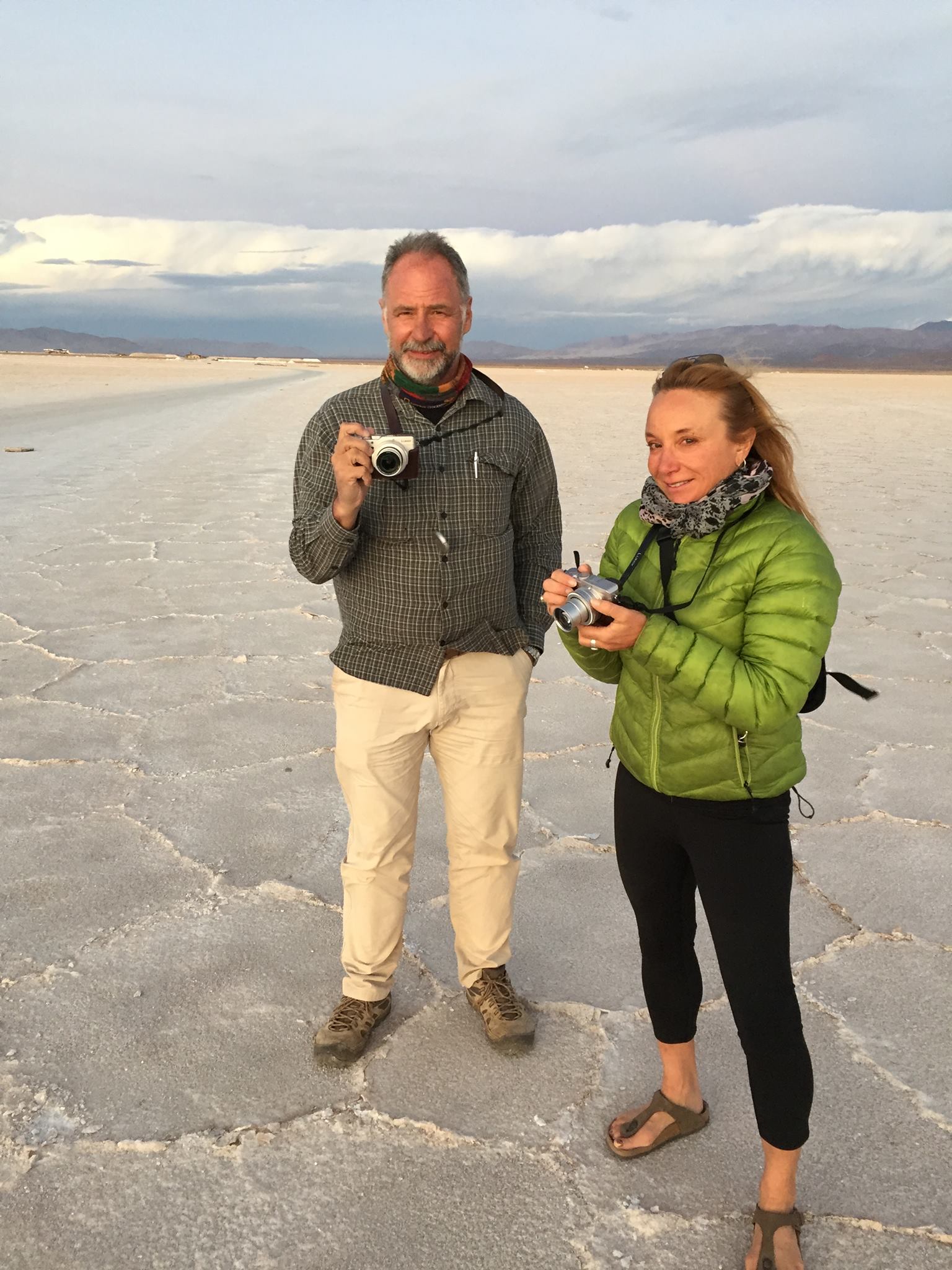 A man holding a camera and a woman in a green jacket stand on a flat, cracked salt plain under a cloudy sky, capturing memories during one of their scenic bike tours.