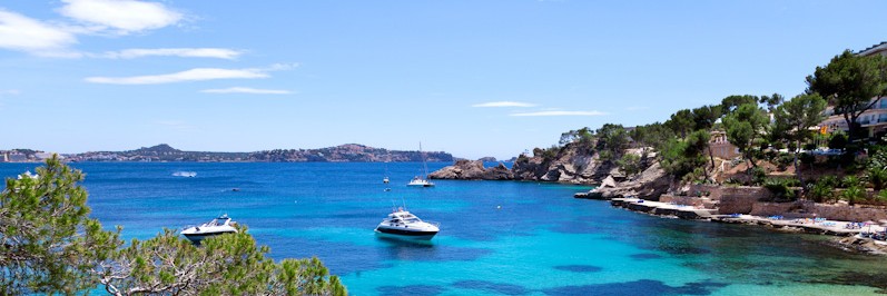 Clear turquoise water with anchored boats near a rocky coastline, bordered by greenery and hills under a blue sky—a perfect view often enjoyed during cycling tours along Italy’s stunning coast.