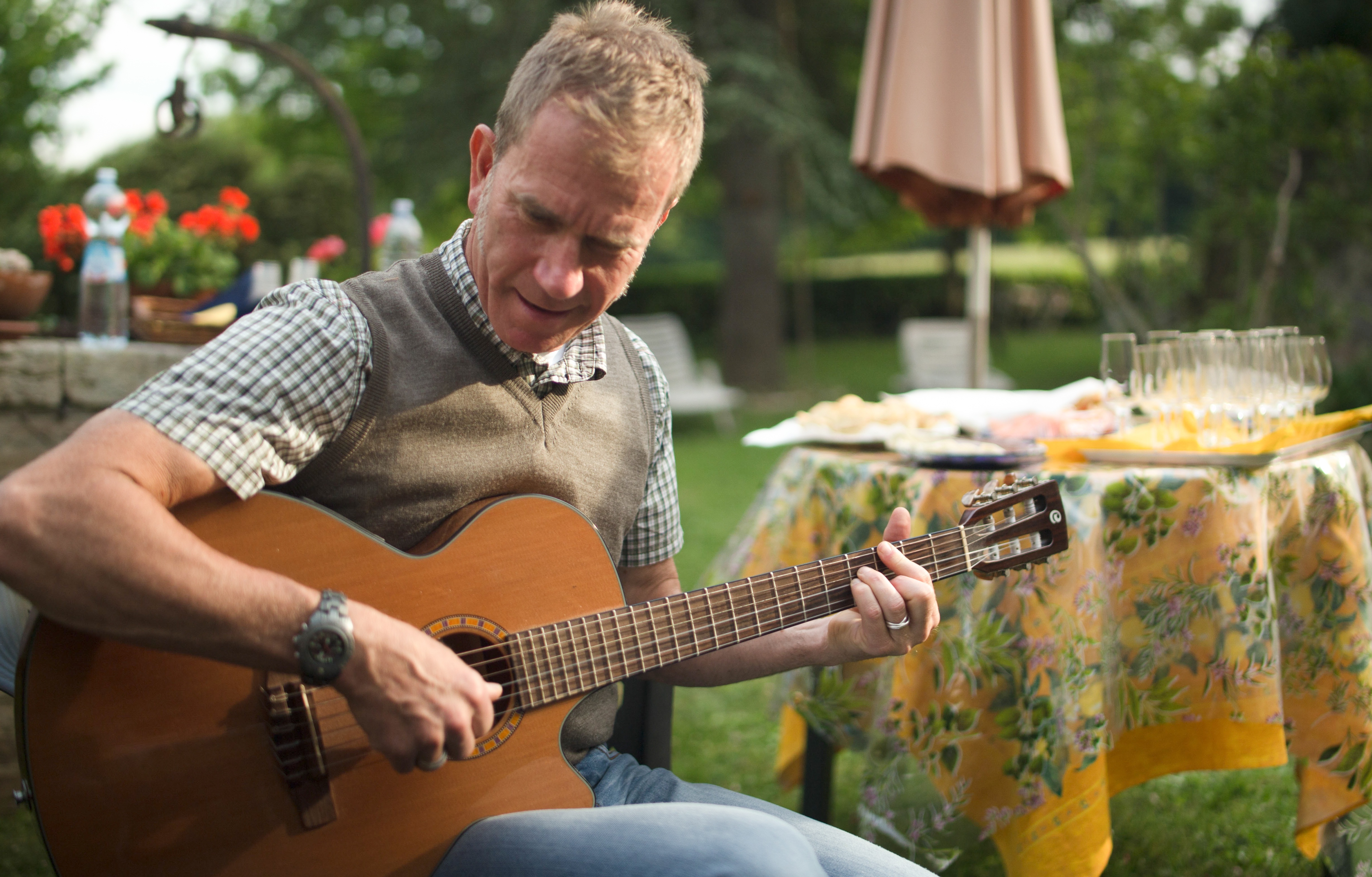 A man sits outdoors playing an acoustic guitar near a table set with glasses and a floral tablecloth, with greenery in the background—capturing the relaxed charm often experienced on scenic bike tours in Italy.