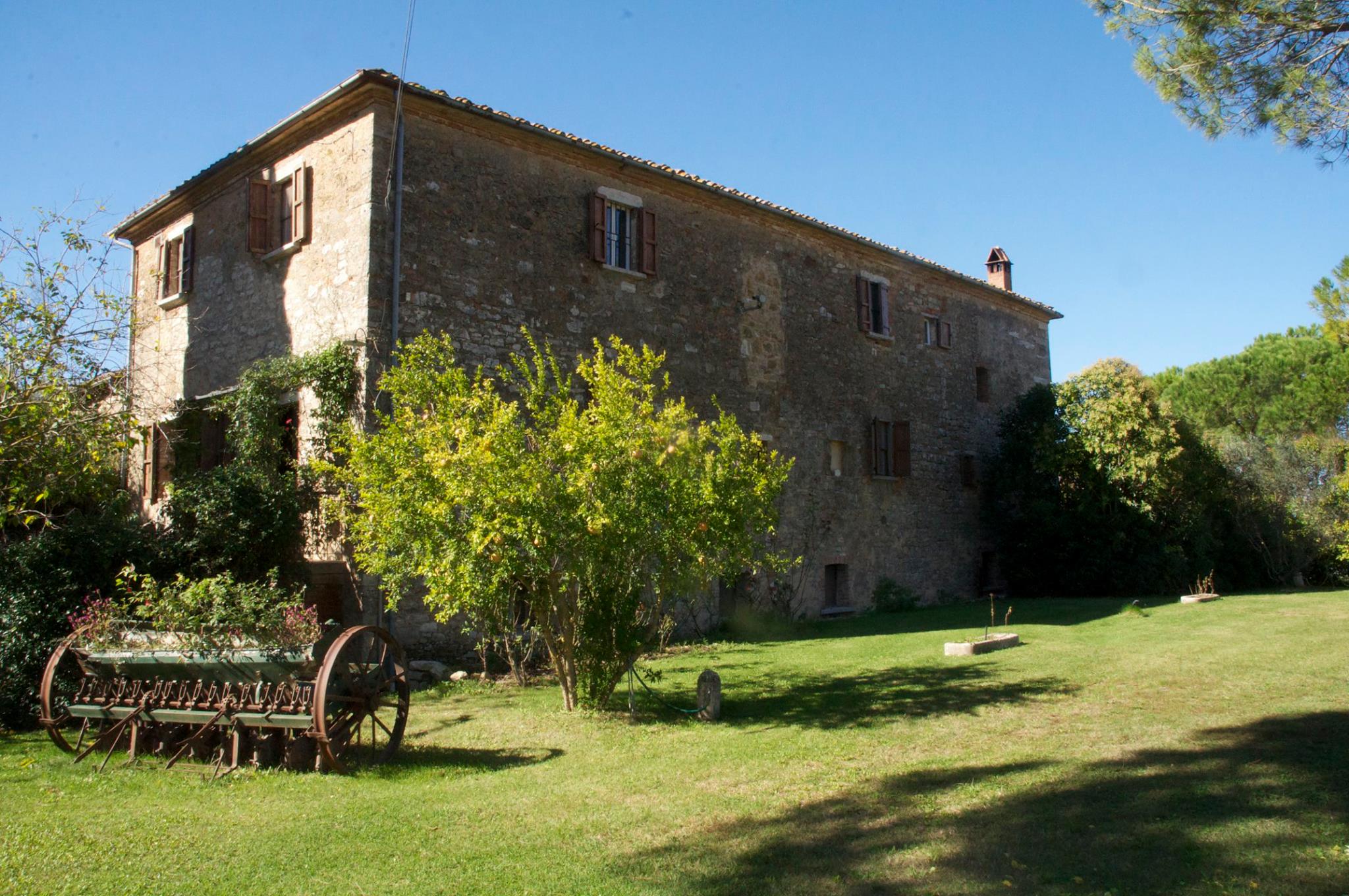 A stone farmhouse with wooden shutters stands on a grassy lawn, surrounded by trees and a vintage metal farming implement in the foreground—an idyllic spot to pause during scenic bike tours through the countryside.