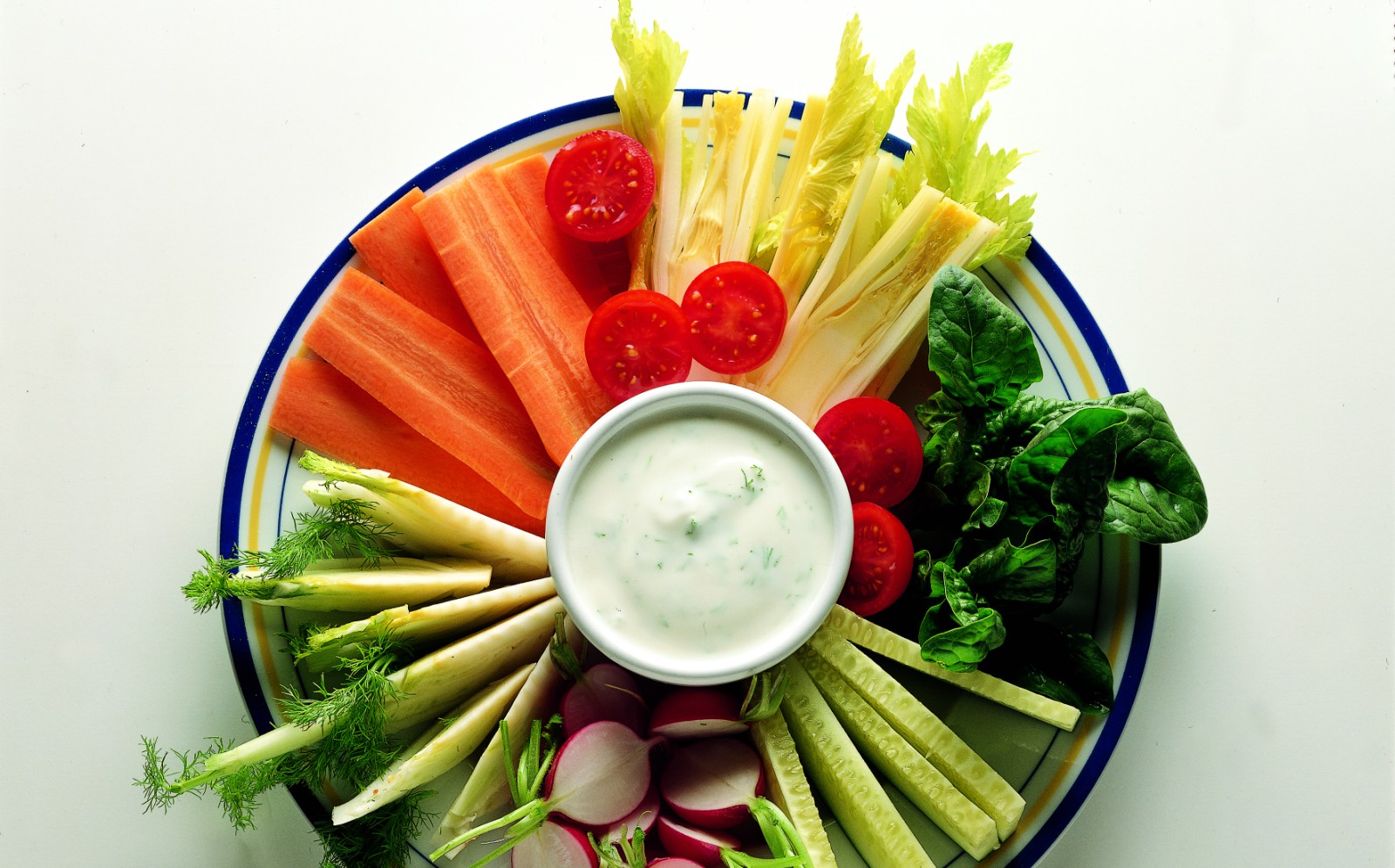 A plate of assorted fresh vegetables including carrots, celery, radishes, tomatoes, fennel, spinach, and cucumber—perfect for enjoying after exhilarating cycling tours or bike tours in Italy—arranged around a bowl of creamy dip.