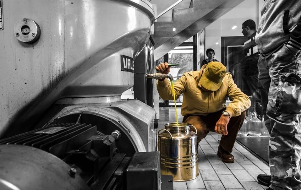 A person in a yellow jacket and cap fills a metal container from a large industrial machine indoors, while others—perhaps part of international bike tours—stand in the background.