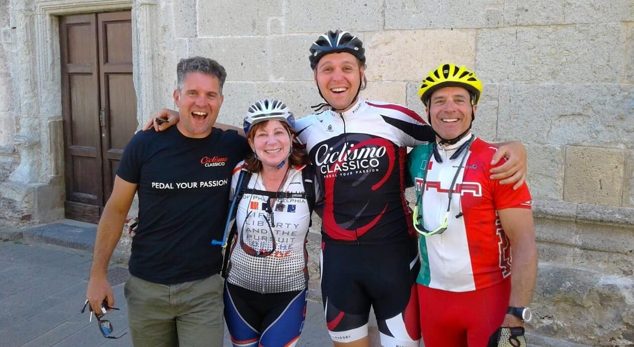 Four smiling adults, three in cycling gear and helmets, stand arm in arm in front of a stone building, posing for a group photo outdoors during one of their scenic bike tours.