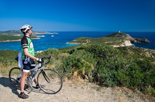 A cyclist in gear stands with a bike on a coastal path, overlooking blue sea, green hills, and a distant lighthouse under a clear sky—an idyllic scene perfect for scenic bike tours.