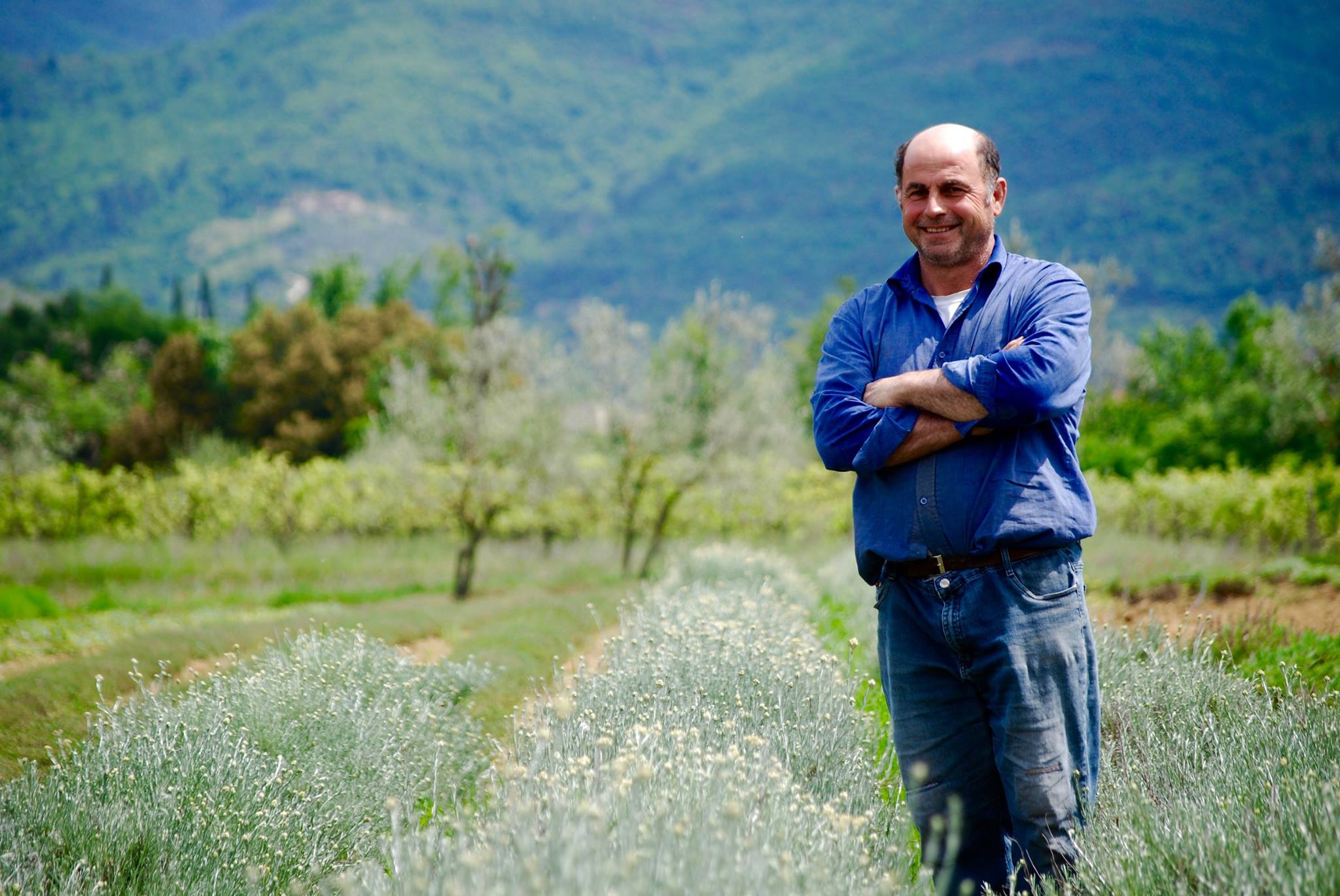Man standing with arms crossed in a field, green hills and trees behind him, wearing a blue shirt and jeans, smiling at the camera—ready to embark on one of the unforgettable bike tours in Italy.