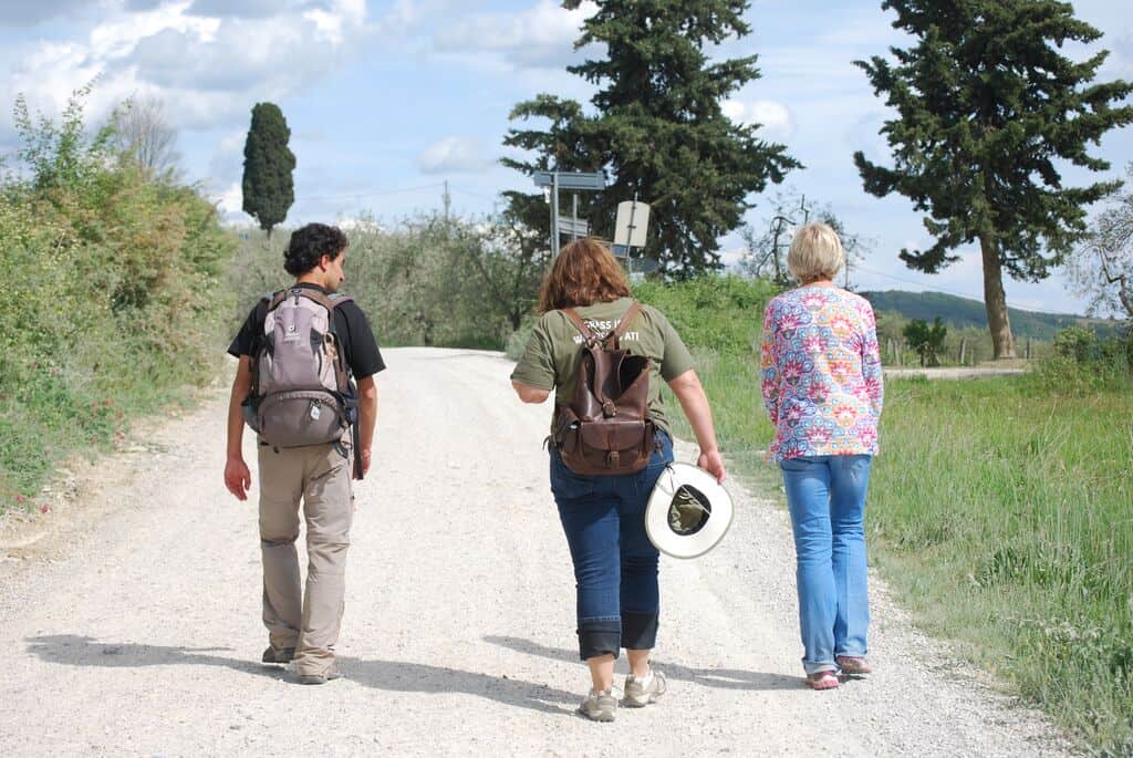 Three people with backpacks walk down a gravel road surrounded by greenery and trees under a partly cloudy sky, reminiscent of the scenic routes enjoyed on bike tours in Italy.
