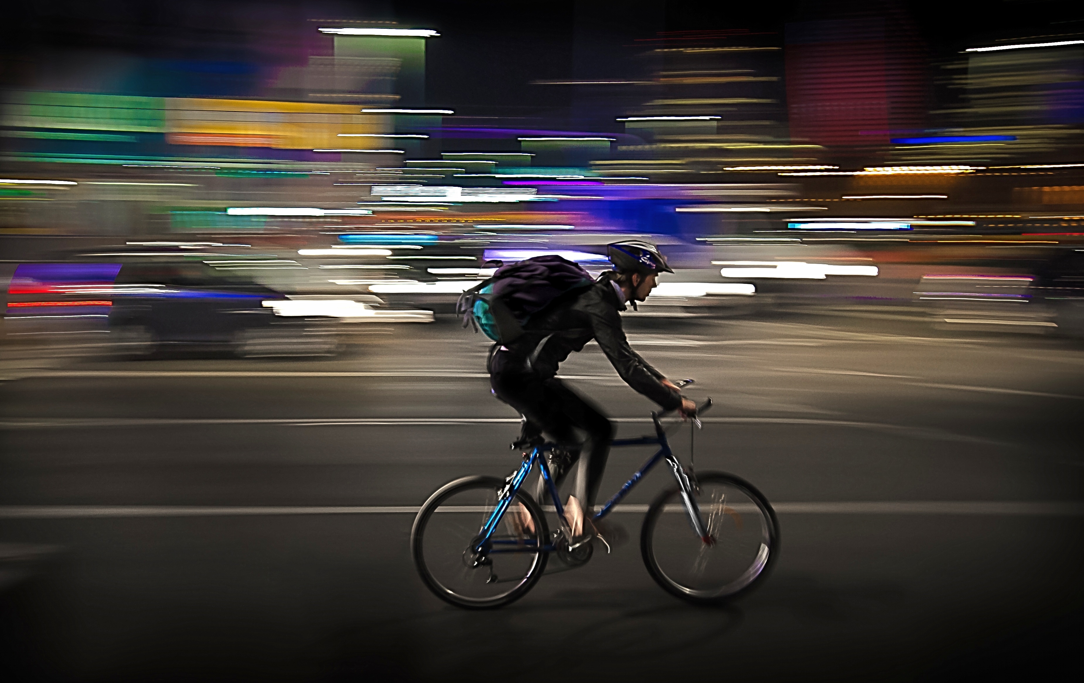 Person wearing a helmet and backpack rides a bicycle through a city street at night, with blurred lights and cars in the background, capturing the excitement of urban cycling tours.