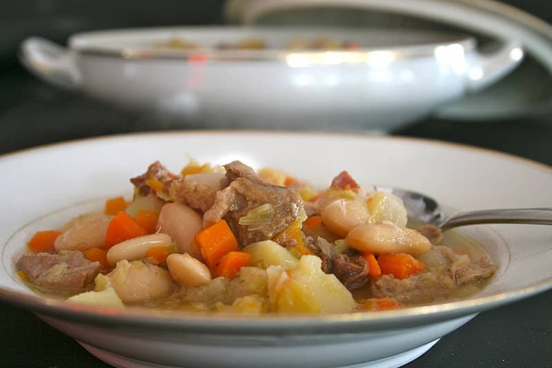 A bowl of hearty stew containing chunks of meat, beans, carrots, and other vegetables rests beside a spoon—the perfect comforting meal after one of your scenic bike tours.