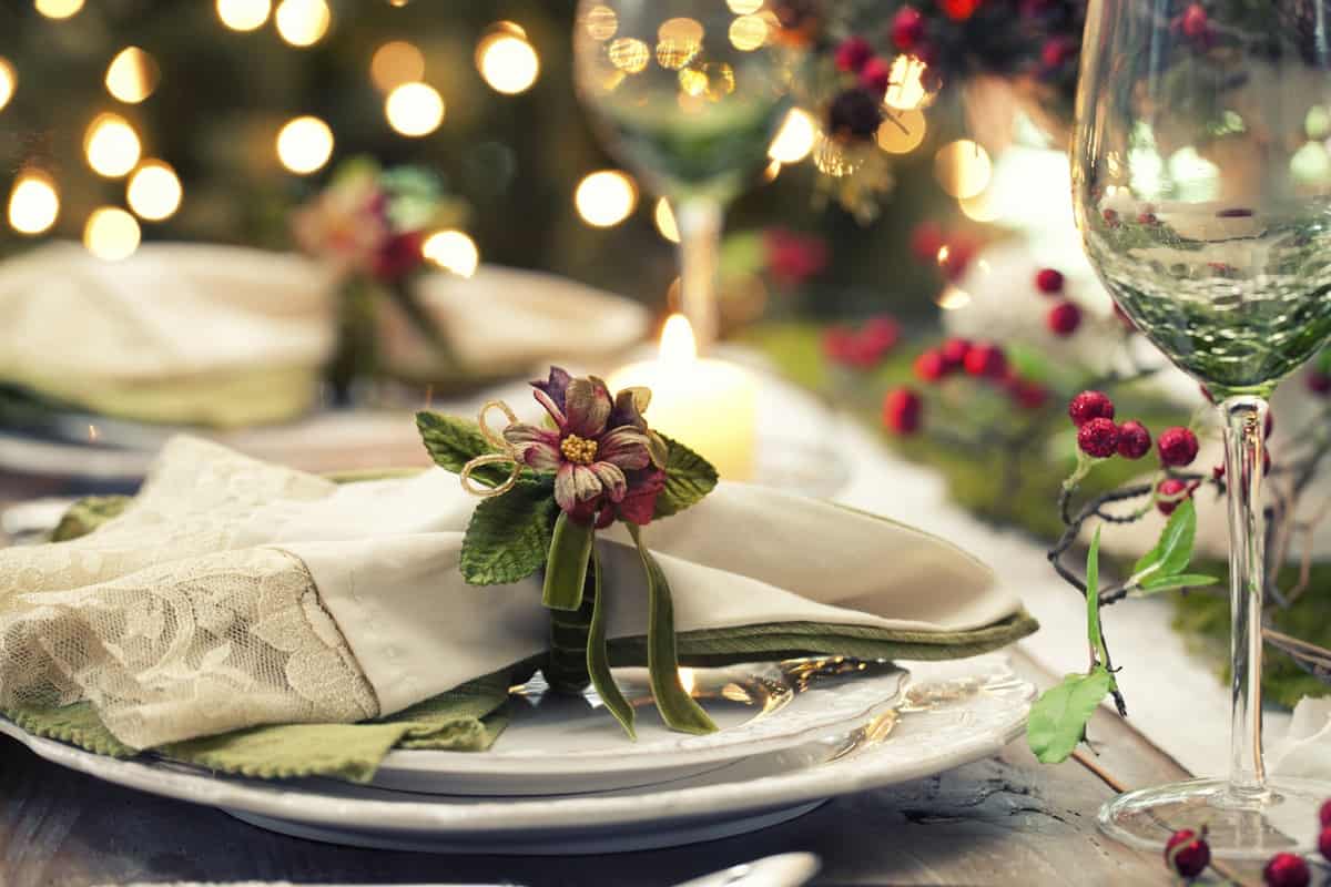 Elegant table setting with a folded napkin, decorative flower, and greenery on a white plate. Perfect for gatherings after scenic bike tours, the scene features a wine glass and festive lights in the background, with red berry accents.