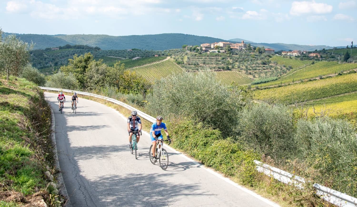 Four cyclists ride along a winding road through green hills and vineyards, enjoying one of the scenic bike tours, with a village visible in the distance under a partly cloudy sky.