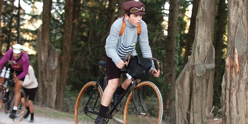 A boy wearing vintage cycling attire rides an old-fashioned bicycle on a forest path, evoking the charm of scenic bike tours, with another cyclist in the background.