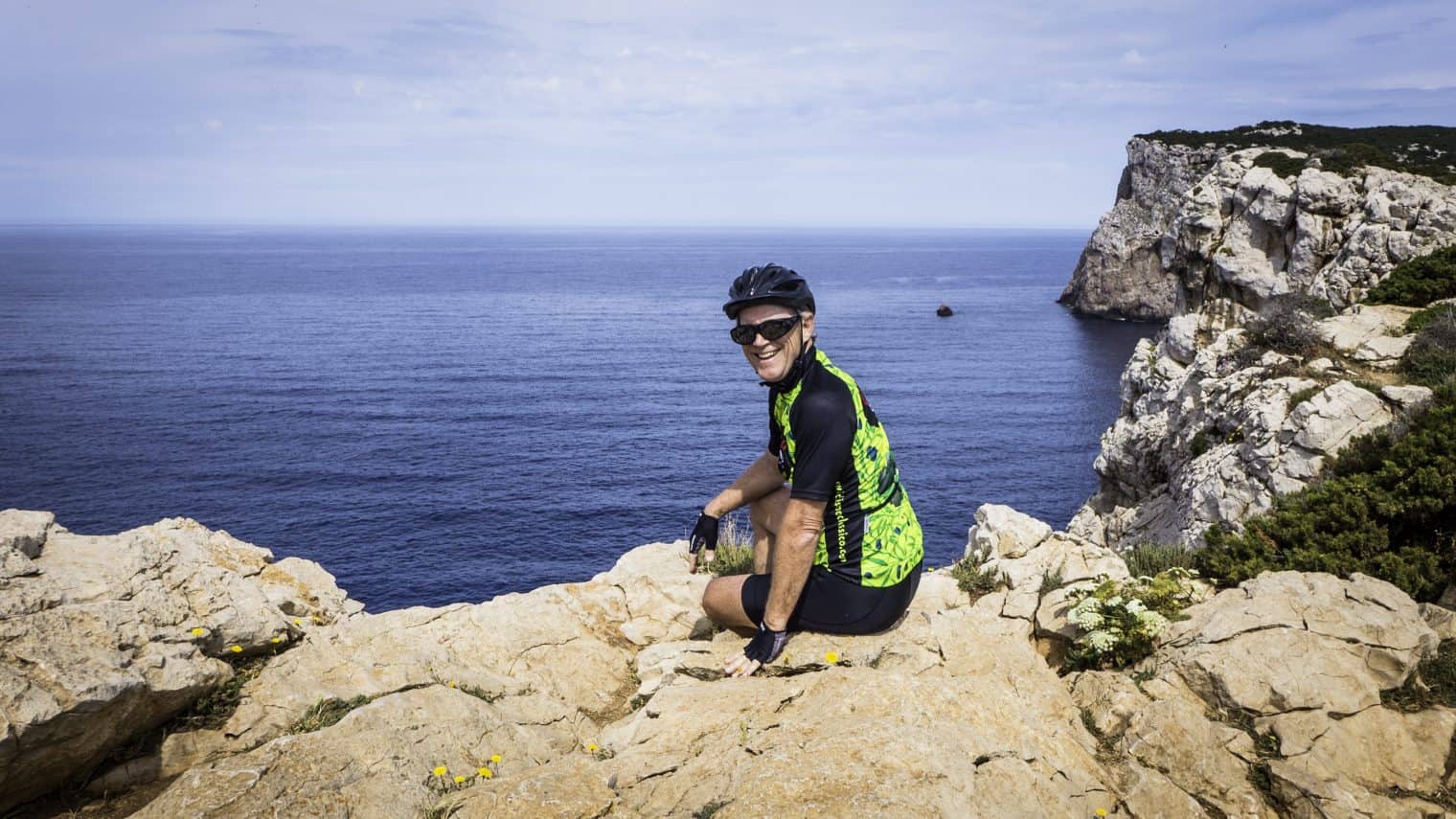 A cyclist in a green and black outfit and helmet sits on a rocky cliff overlooking the blue sea, taking in the stunning views—a perfect moment from scenic bike tours. A large rock formation rises majestically in the background.