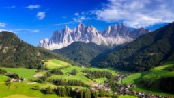 A small village sits in a green valley, surrounded by forests and mountains under a blue sky with scattered clouds—a perfect scene for bike tours in Italy.