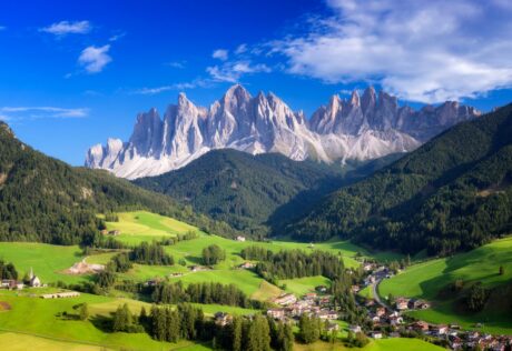 A small village sits in a green valley, surrounded by forests and mountains under a blue sky with scattered clouds—a perfect scene for bike tours in Italy.