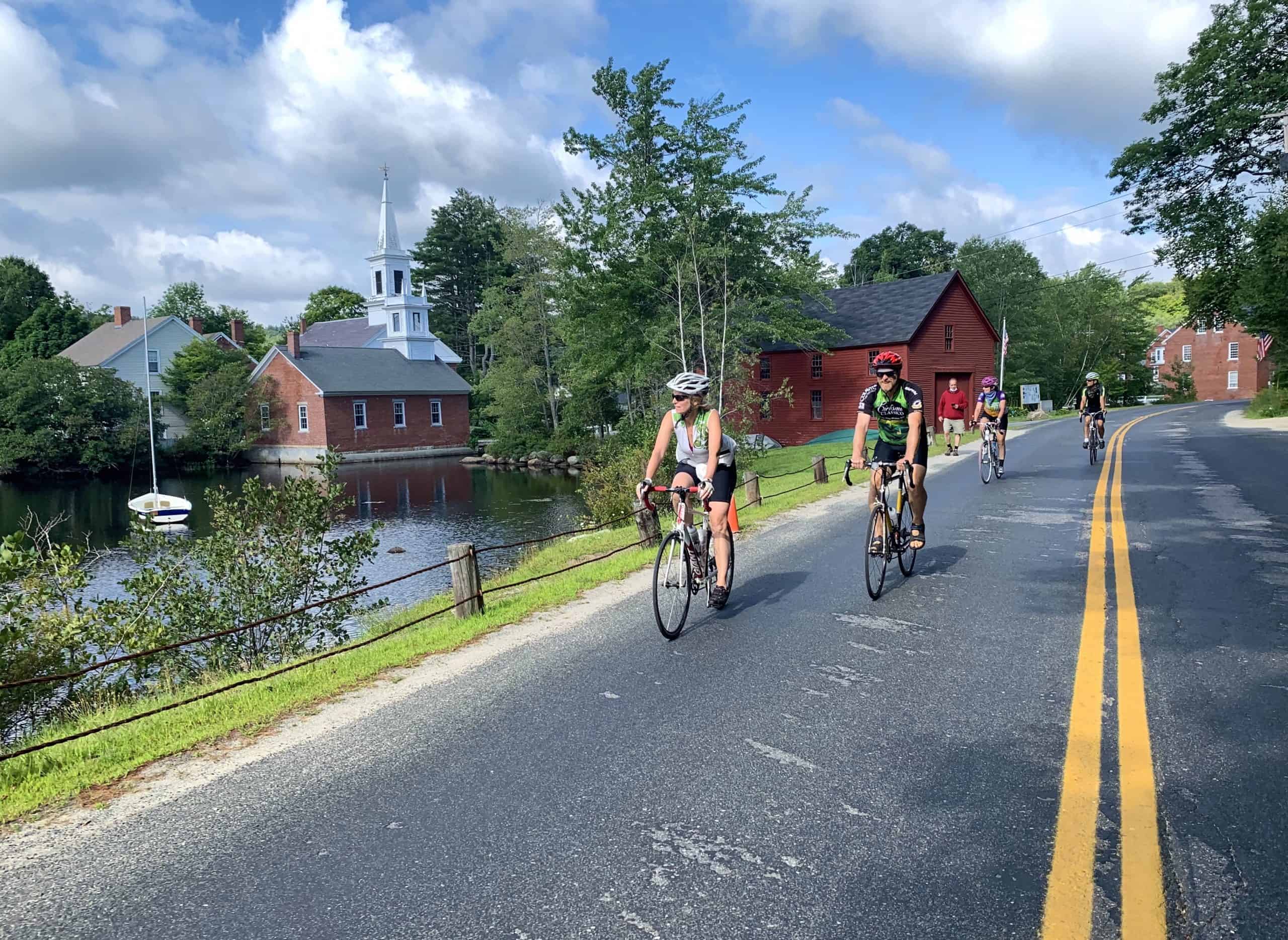 Four cyclists ride on a rural road beside a pond, passing red buildings and a white church steeple beneath a partly cloudy sky—a perfect setting for scenic bike tours through charming countryside.