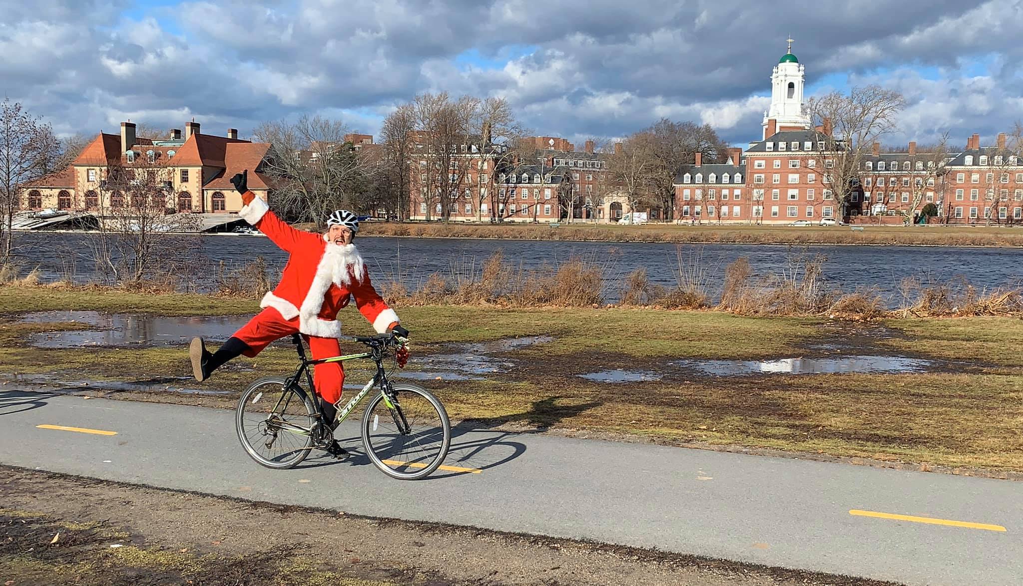 Dressed as Santa Claus, a person rides one-handed along a riverside path, passing historic brick buildings beneath a cloudy sky—capturing the festive spirit you might find on scenic bike tours.