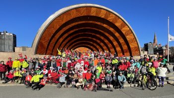 A large group of cyclists in colorful attire poses in front of the Hatch Memorial Shell on a sunny day, capturing the spirit of scenic bike tours, with blue sky and flags in the background.