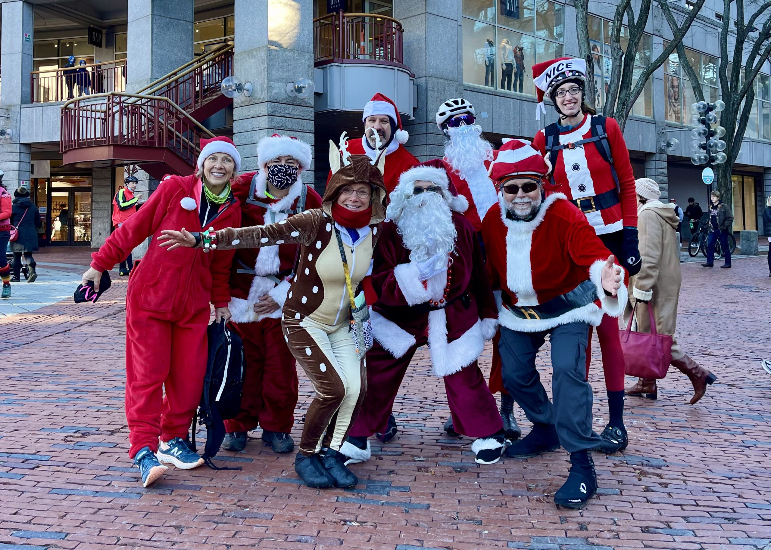 A group of people in festive costumes, including Santa suits and holiday-themed outfits, pose together and smile on a brick-paved street during one of the lively Italy bike tours in an urban area.