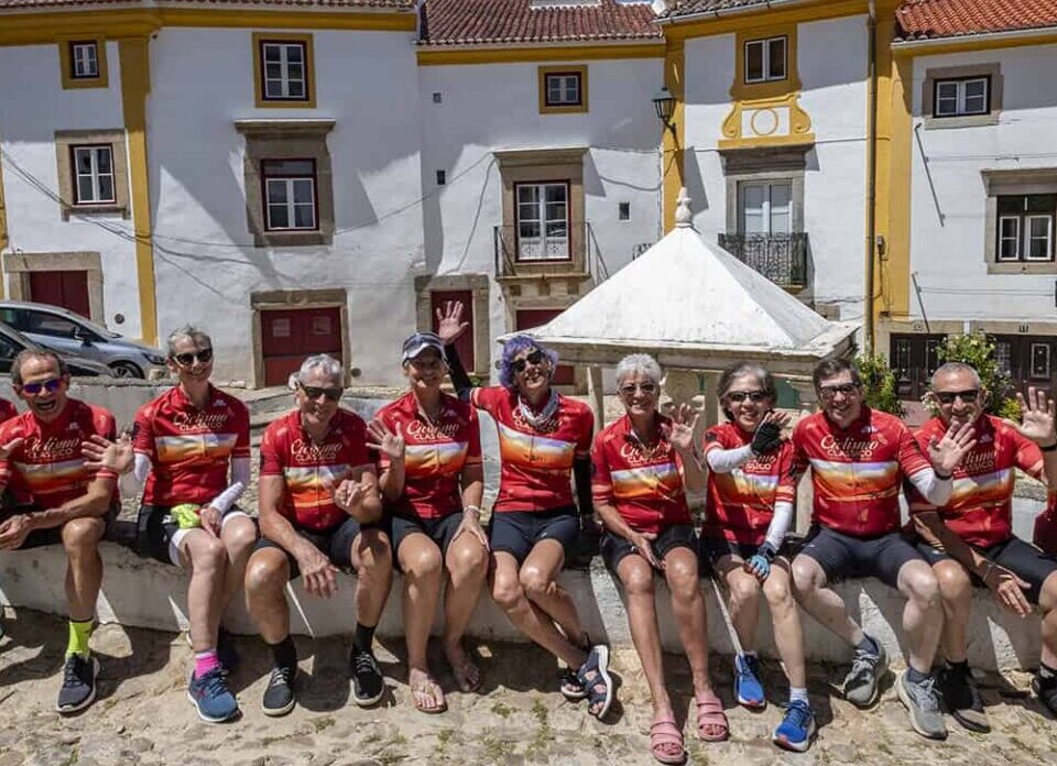 Group of cyclists wearing matching red jerseys resting on a stone bench in a scenic Italian village, enjoying Italy bike tours with Ciclismo Classico.