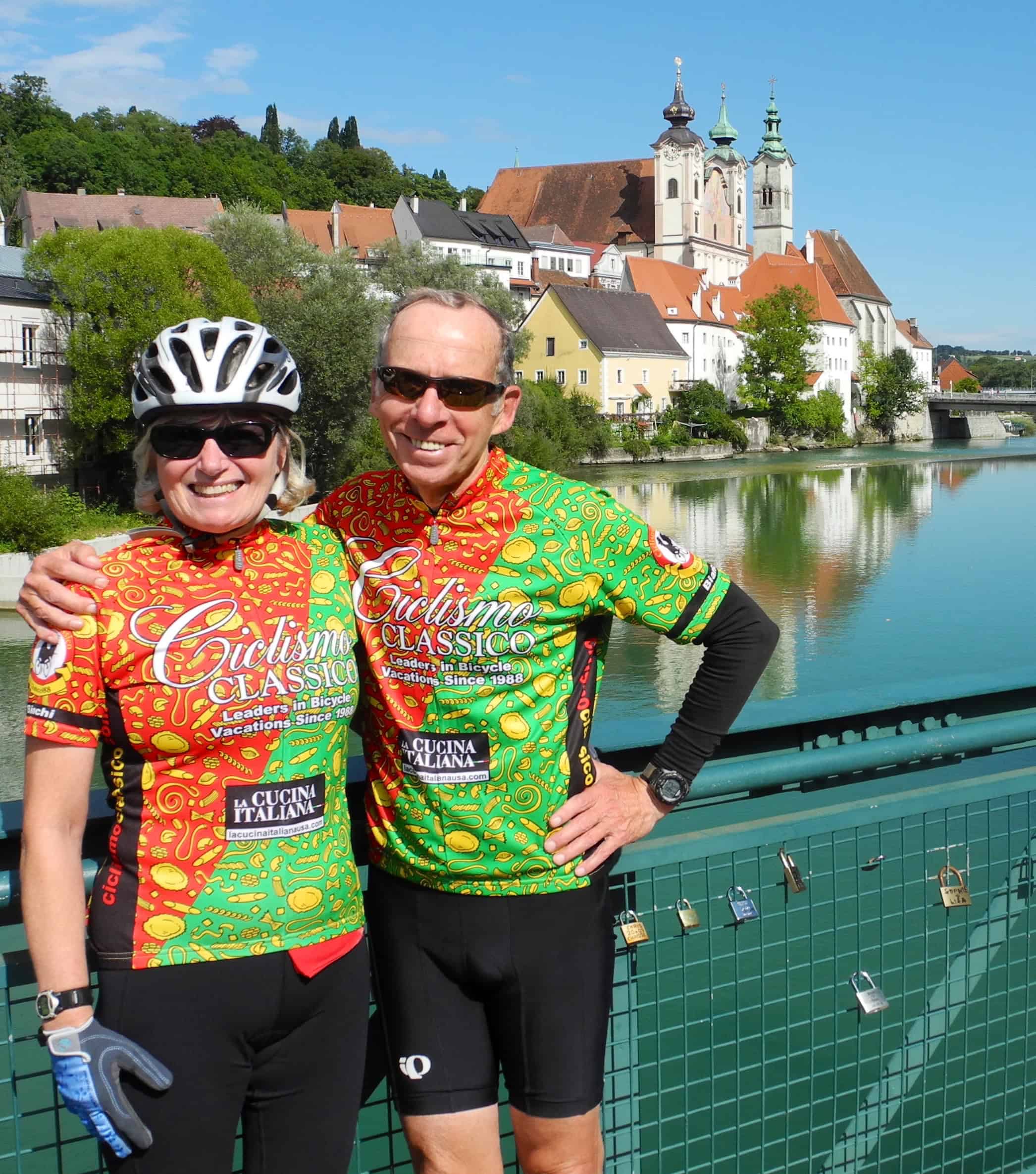 Colorful Italian cycling jerseys worn by a smiling senior couple during a bike tour in Italy, overlooking a historic town and river, on a bright sunny day.