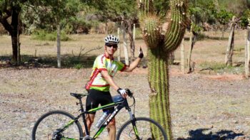Vibrant cyclist in colorful gear pointing at a tall cactus during an Italy bike tour in a desert landscape.