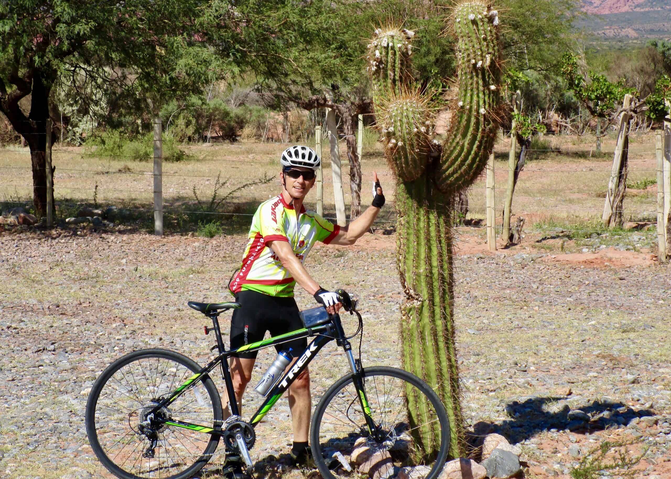 Vibrant cyclist in colorful gear pointing at a tall cactus during an Italy bike tour in a desert landscape.