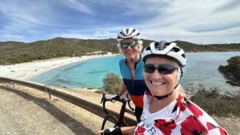 A sunny day cycling tour along a scenic Italian coastline with smiling women in helmets and sunglasses, enjoying the picturesque views of turquoise waters and lush hills.