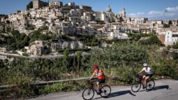 Vibrant cyclists enjoying Italy's scenic bike tour along a hillside road with a historic town in the background during daytime.