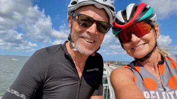 Vibrant image of smiling cyclists in safety helmets and sunglasses enjoying a scenic bike ride along a waterfront in Italy under a blue sky.