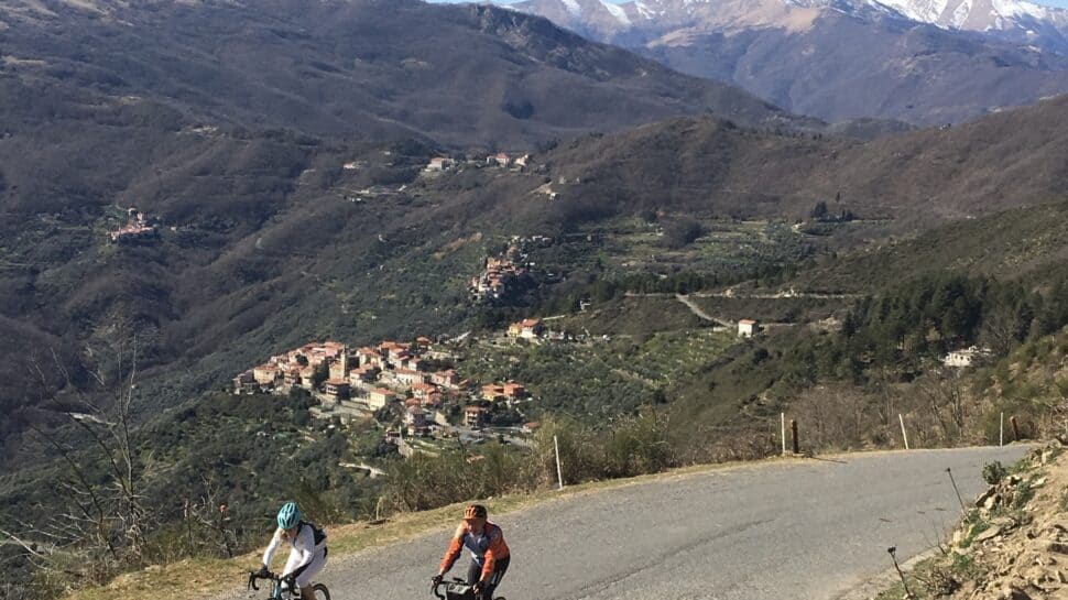 Bikers riding mountain bikes on a scenic Italy mountain road with village and snow-capped peaks in the background.