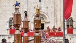 Colorful Italian festival with dozens of spectators, historical costumes, and religious statues outside a classic stone church in Italy.