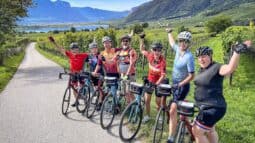 Sunny group cycling tour through Italian vineyards with scenic mountain views and a lake in the background, showcasing Italy bike tours by Ciclismo Classico.