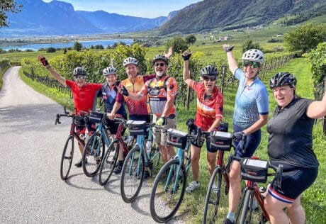 Sunny group cycling tour through Italian vineyards with scenic mountain views and a lake in the background, showcasing Italy bike tours by Ciclismo Classico.