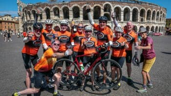 Enjoying a group cycling tour in front of the Roman Arena in Verona, Italy, with a team of cyclists in orange jerseys celebrating their ride.