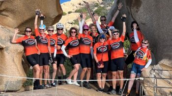 A group of cyclists in matching orange jerseys and helmets posing together on a scenic rocky coastal trail in Italy, celebrating their bike tour adventure with the sea in the background.