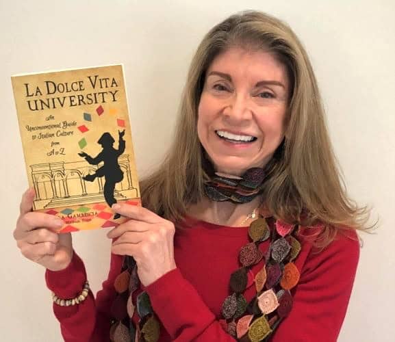 Woman holding a book titled "La Dolce Vita University" smiling at the camera, promoting Italian culture and lifestyle, ideal for Italy bike tours and exploring Italy's scenic beauty.