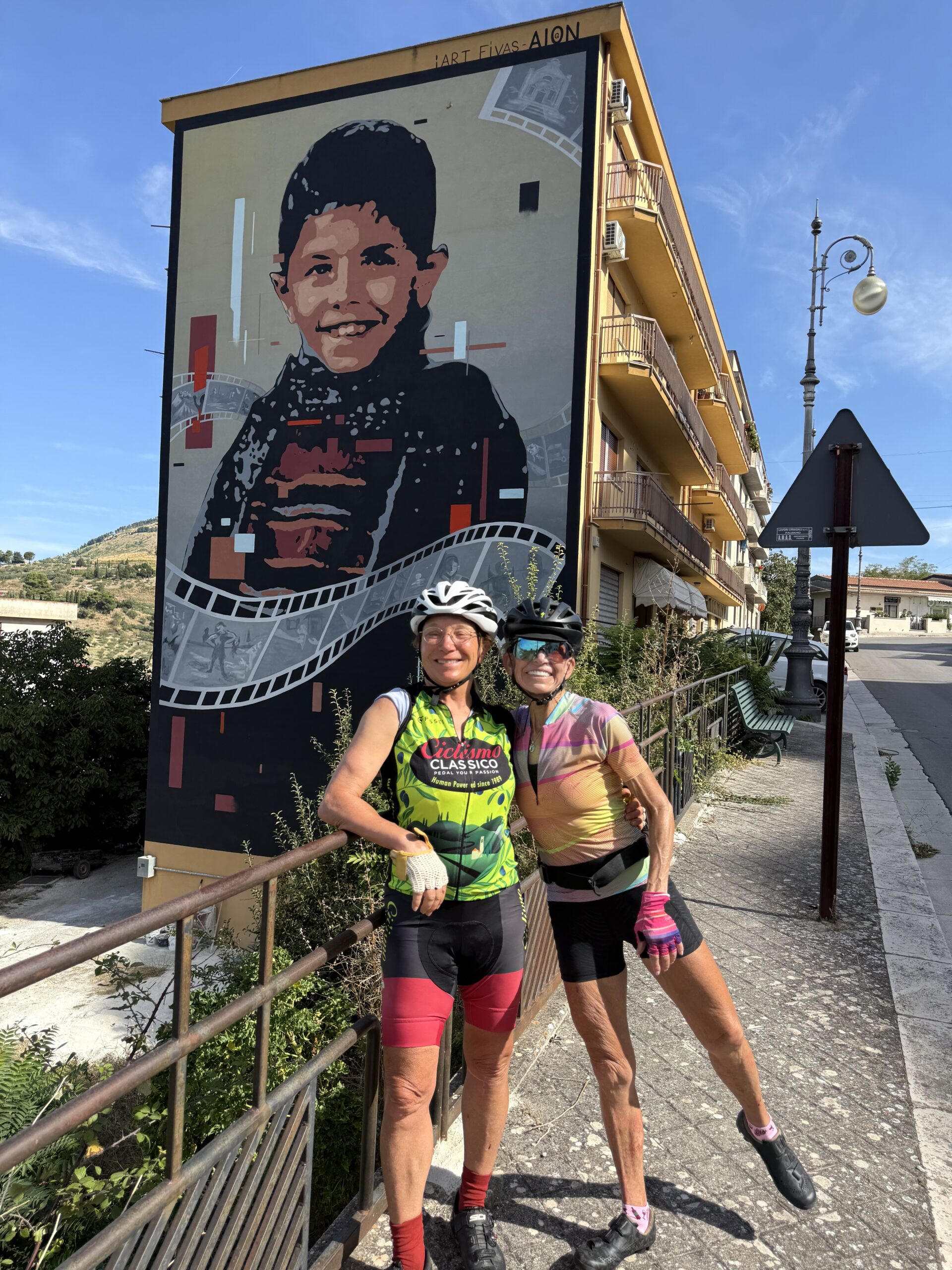Two women in cycling gear standing in front of a large mural of a smiling boy, promoting Italy bike tours with Ciclismo Classico.