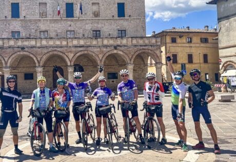Group of cyclists in colorful gear standing with bikes in a historic Italian town square, showcasing scenic bike tours in Italy for cycling enthusiasts.