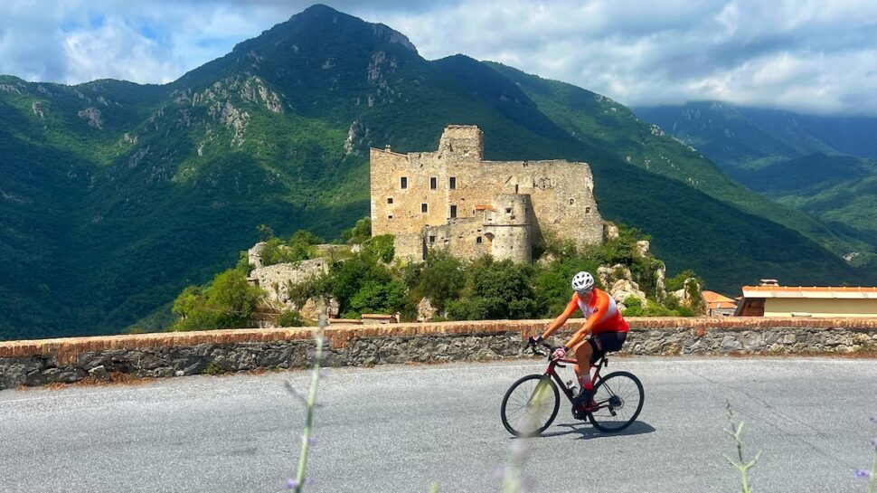 Cyclist riding a bike on a mountain road with a historic castle and lush green mountains in Italy, perfect for bike tour enthusiasts.