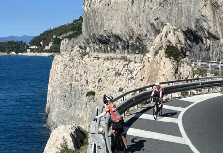 Cyclists riding along a scenic coastal road in Italy with cliffs and blue sea in the background, showcasing Italy bike tours and scenic cycling routes.