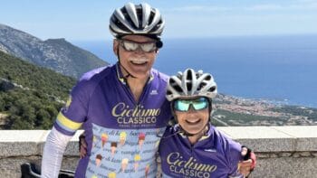 Two cyclists in purple jerseys smiling with scenic coastal view in Italy, promoting bike tours and cycling experiences in Italy by Ciclismo Classico.