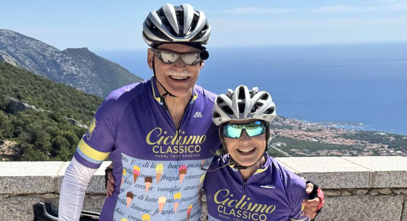 Two cyclists in purple jerseys smiling with scenic coastal view in Italy, promoting bike tours and cycling experiences in Italy by Ciclismo Classico.